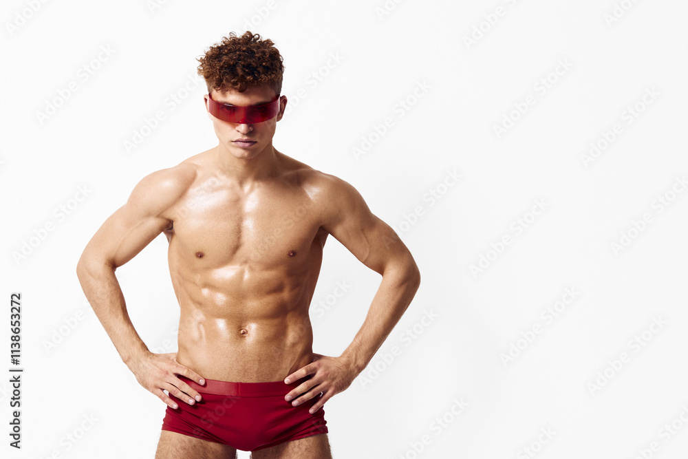 Fototapeta premium Confident athletic man in red swimwear posing against a plain white background, showcasing fitness and body positivity, exuding strength and self-assurance.