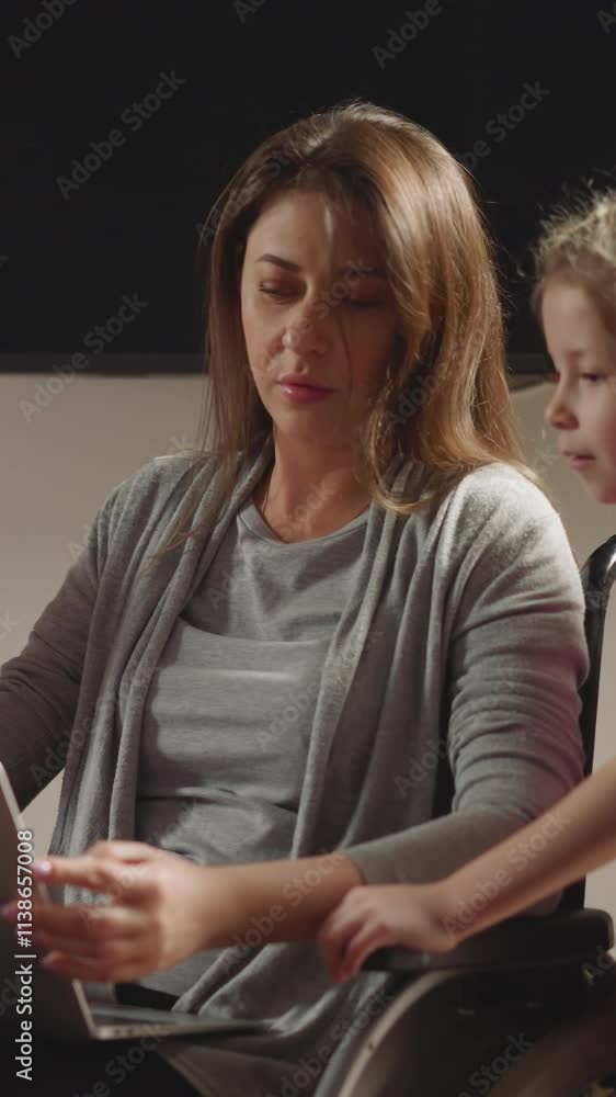 Curious preschooler and toddler bother mom working on laptop. Cute kids watch mother with paralysis using modern gadget in wheelchair at home Curious preschooler and toddler bother mom working on laptop. Cute kids watch mother with paralysis using modern gadget in wheelchair at home