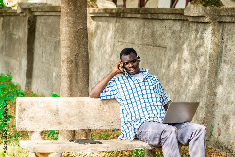 Fototapeta premium Young cheerful black man using mobile phone and laptop computer. Modern lifestyle