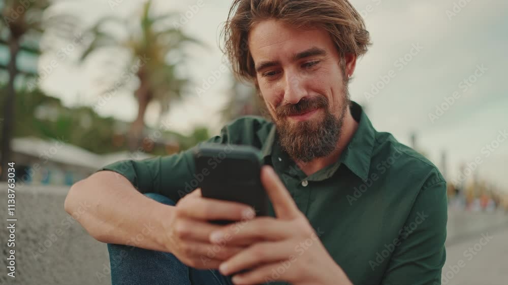 Close-up portrait of smiling man with a beard chatting on the embankment background. Frontal close-up of happy young male hipster using mobile phone Close-up portrait of smiling man with a beard chatting on the embankment background. Frontal close-up of happy young male hipster using mobile phone