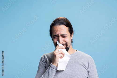 Sad melancholic person with anxiety crying and whining on camera, feeling distraught and in an emotional crisis against blue background. Young guy wiping his tears with a tissue. photo