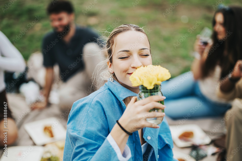 Fototapeta premium A young woman closes her eyes to savor the scent of yellow flowers during a picnic with friends. The setting is relaxed, capturing a joyful outdoor experience and a moment of contentment.