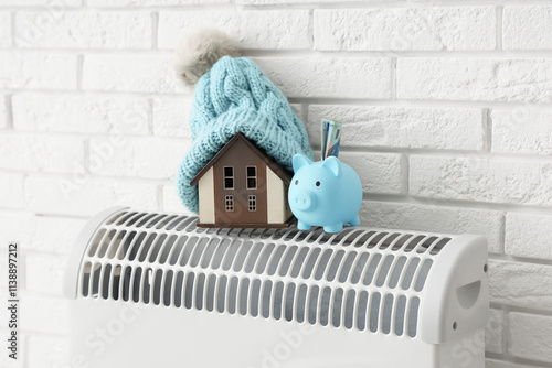 Piggy bank with money, house model, and warm hat on convector heater near white brick wall. Heating saving concept photo