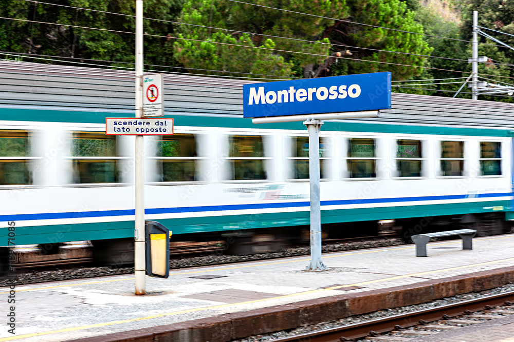 A train speeds through Monterosso station, located in Cinque Terre, Italy. The vibrant colors of the train contrast against the green landscape and platform at dusk.