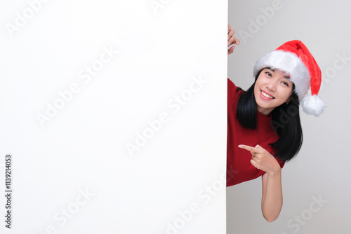 A woman wearing santa christmas hat peeking out behind wall and pointing on it with happy face photo