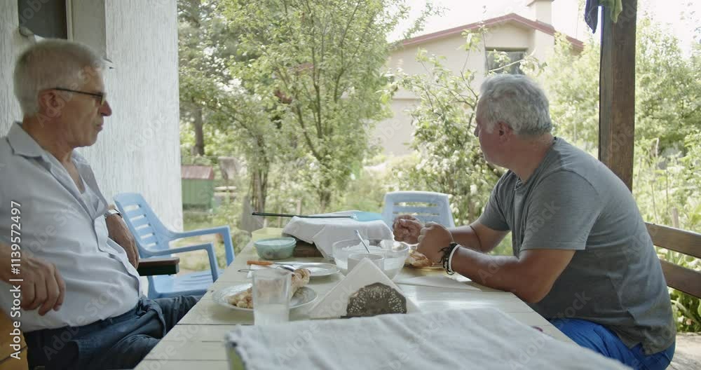Two older men share a meal and conversation on their porch, enjoying retirement and the summer weather. Two older men share a meal and conversation on their porch, enjoying retirement and the summer weather.