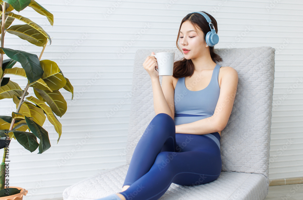 Fototapeta premium Asian young beautiful cheerful woman sporty active energetic girl in exercise sportswear and headphones listen music drink coffee in cup sitting relax on cozy sofa in living room lifestyle at home.