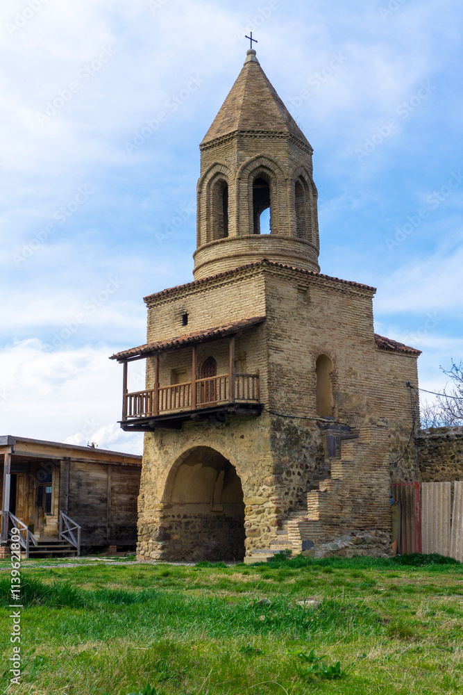 Fototapeta premium Bell tower with wooden balcony and entrance to the courtyard of Samtavisi Cathedral. Green grass, bright blue sky with clouds
