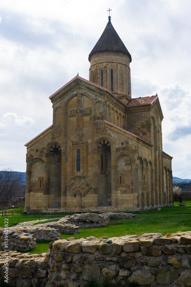 Samtavisi Cathedral in a courtyard with green grass. Stone walls with frescoes, orange tiled roof. Stone paths Samtavisi Cathedral in a courtyard with green grass. Stone walls with frescoes, orange tiled roof. Stone paths