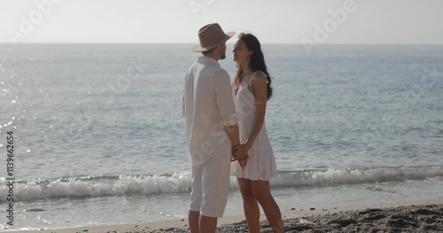 A cheerful couple enjoys a romantic walk along the beach, holding hands and laughing. The serene ocean, soft sand, and natural rock formations create a perfect backdrop for love and happiness.