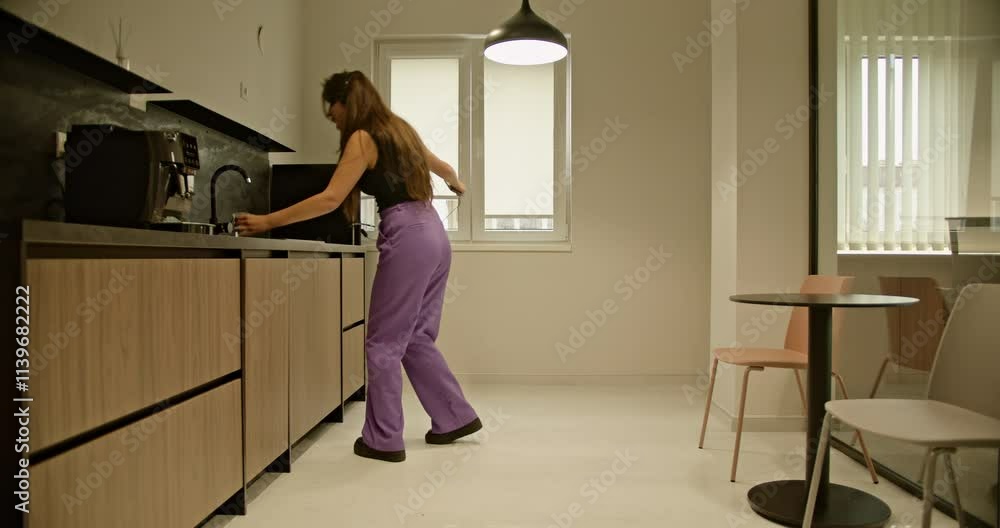 A young woman dances happily in the office kitchen, enjoying a break and listening to music on her headphones. A young woman dances happily in the office kitchen, enjoying a break and listening to music on her headphones.