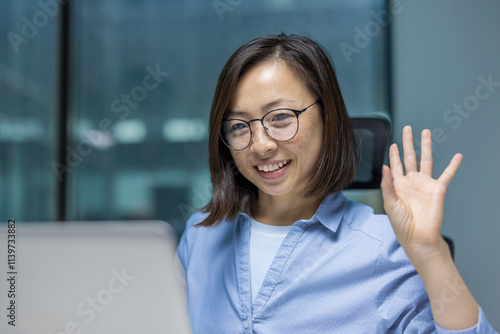 Free Download Asian businesswoman smiling and waving engaging in a video conference call. She is seated at her office desk wearing glasses and a professional shirt, creating a welcoming and cheerful atmosphere - GFXLoad