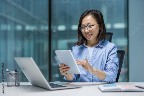 Asian woman dressed professionally, using a tablet at her desk in office. She appears focused and engaged, demonstrating concepts of technology usage and productivity in professional environments.