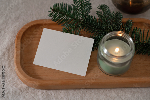 Blank paper card mockup, green candle with soft dim light, fir branch on wooden tray on table, cozy aesthetic winter home still life, template for brand business card, postcard or invitation photo