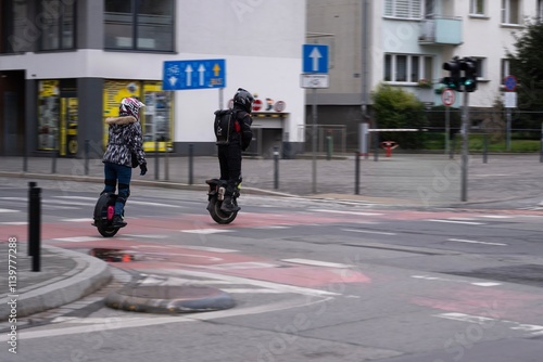 Two people riding electric unicycles across a city street, wearing helmets and protective gear photo