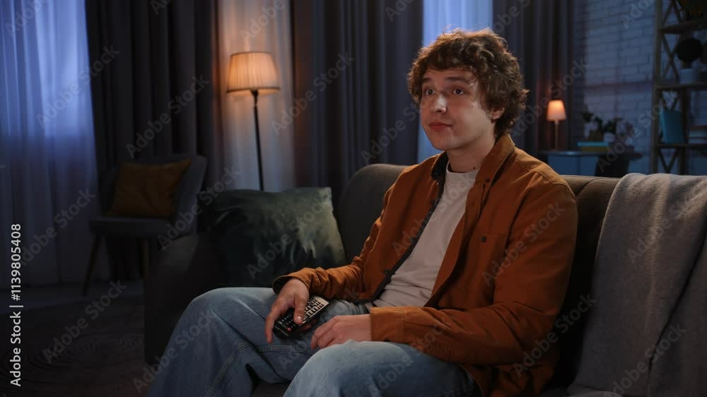 Young man spending evening at home sitting on sofa in living room, switching tv with remote watching program, looks surprised and watching video with interest. Young man spending evening at home sitting on sofa in living room, switching tv with remote watching program, looks surprised and watching video with interest.