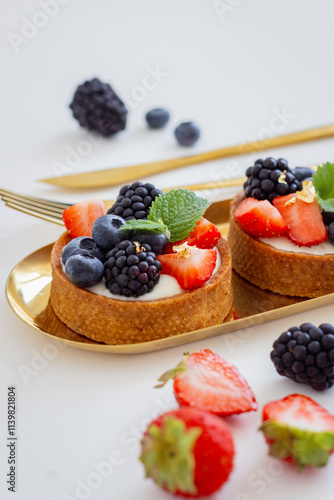Tartlets with Fresh Blueberries, Strawberries, and Blackberries on a Gold Stand with Golden Cutlery on a White Background