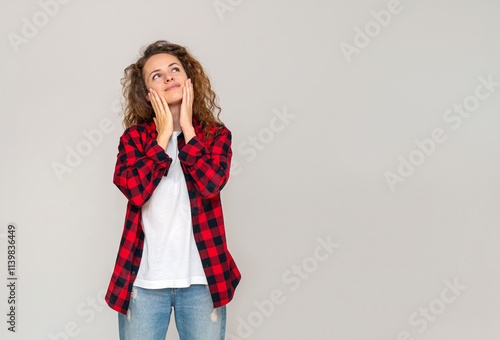 Woman wearing red shirt standing next to a grey background touching face with her hands, looking up and smiling.