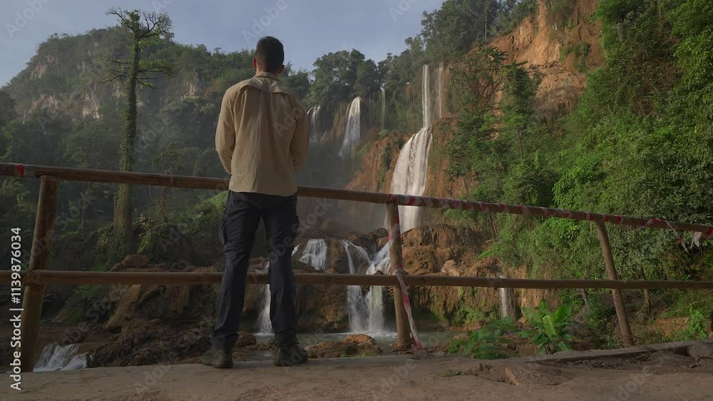 A man walks into frame to admire Thailand's largest waterfall before turning and walking out of frame. A man walks into frame to admire Thailand's largest waterfall before turning and walking out of frame.