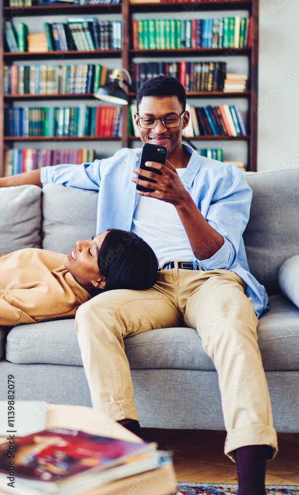 © BullRun - African American man in classic eyewear enjoying cellphone messaging on modern cellular technology spending weekend with lovely wife lying on knees and reading literature book, leisure time