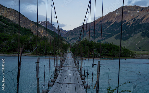 A rustic suspension bridge stretches across a serene river, framed by rugged mountain peaks in the distance. The wooden planks and simple design evoke a sense of adventure, while the calm water below  photo