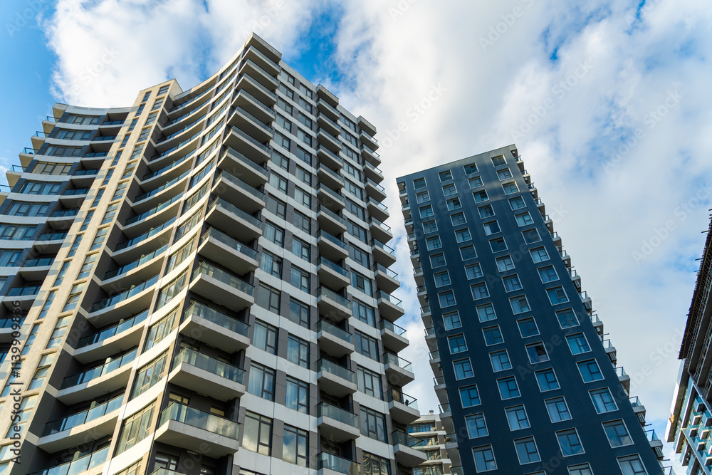Fototapeta premium Modern apartment buildings with glass balconies standing tall under a blue sky with white clouds, showcasing contemporary urban architecture