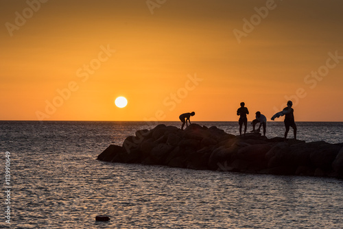Fototapeta Naklejka Na Ścianę i Meble -  Group of youths enjoy a peaceful sunset moment on a rocky outcrop on a beach in Noord, along the coast of Aruba