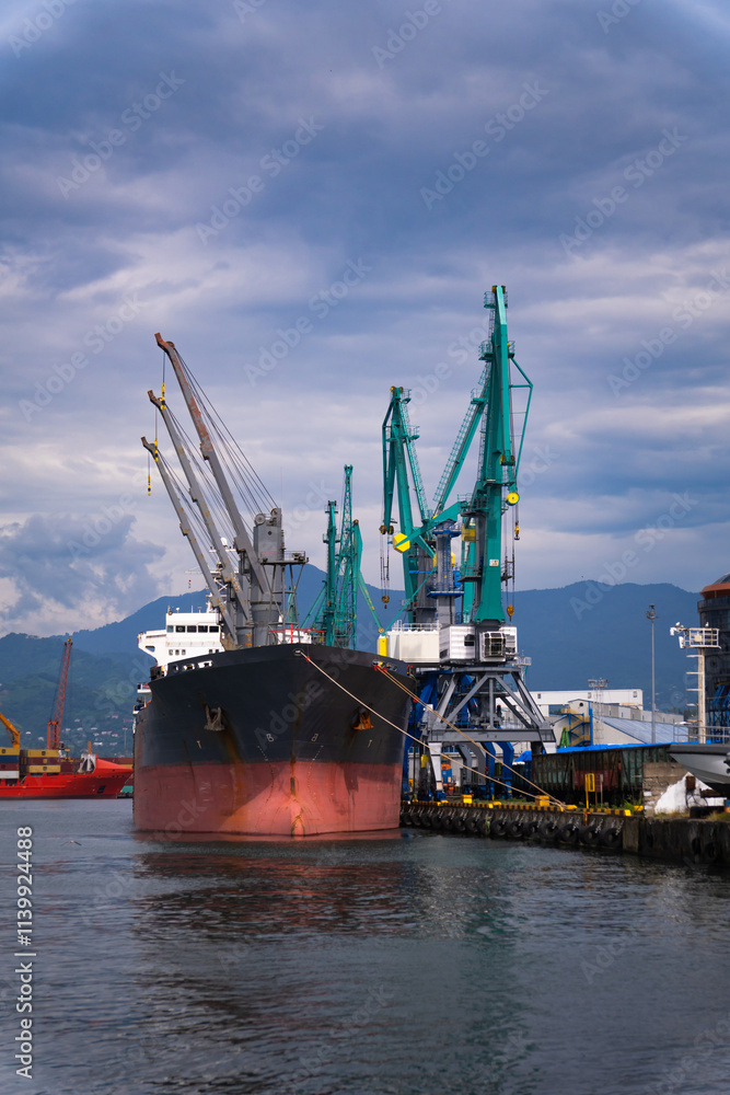 Large cargo ship is docked at a commercial port, being loaded by cranes under a cloudy sky. The ship is being prepared for its next voyage, transporting goods across the sea Large cargo ship is docked at a commercial port, being loaded by cranes under a cloudy sky. The ship is being prepared for its next voyage, transporting goods across the sea