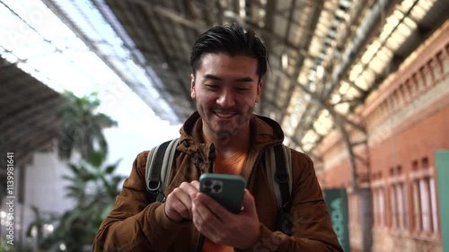 Young asian man with backpack is using smartphone and smiling while standing in atocha railway station in madrid, spain, enjoying his travel or vacation