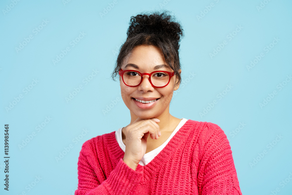 Smiling young woman with red glasses and pink sweater touching chin on blue background Smiling young woman with red glasses and pink sweater touching chin on blue background