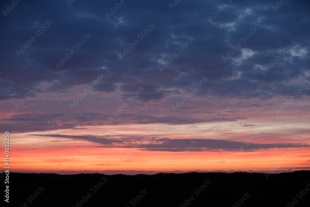 Sunset hues paint a dramatic sky above dark silhouettes of hills. Clouds transition from dark to vibrant orange-pink. Sunset hues paint a dramatic sky above dark silhouettes of hills. Clouds transition from dark to vibrant orange-pink.