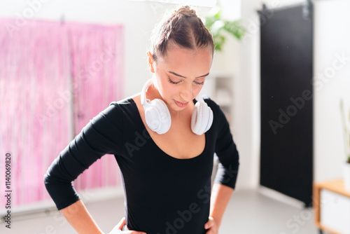 A close-up image of a ballerina in a black leotard with white headphones around her neck, standing gracefully in a bright studio with pink curtains softly blurred in the background. photo