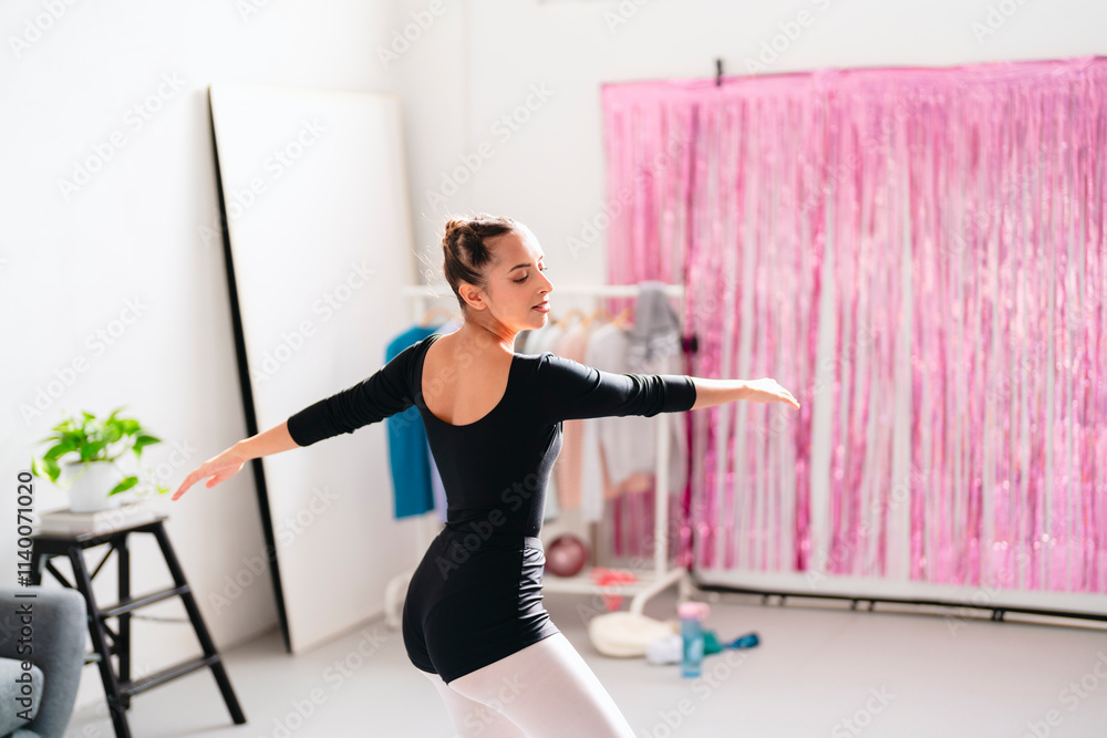 A ballerina dressed in black leotard and white tights poses elegantly in a bright studio with a modern gray couch, a clothing rack, and decorative pink streamers in the background. A ballerina dressed in black leotard and white tights poses elegantly in a bright studio with a modern gray couch, a clothing rack, and decorative pink streamers in the background.