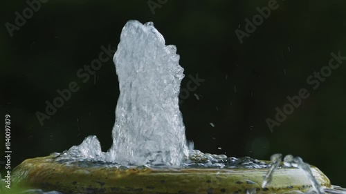 Flow of clean water is splashing out upwards from a small artificial water fountain with blurry natural background. photo