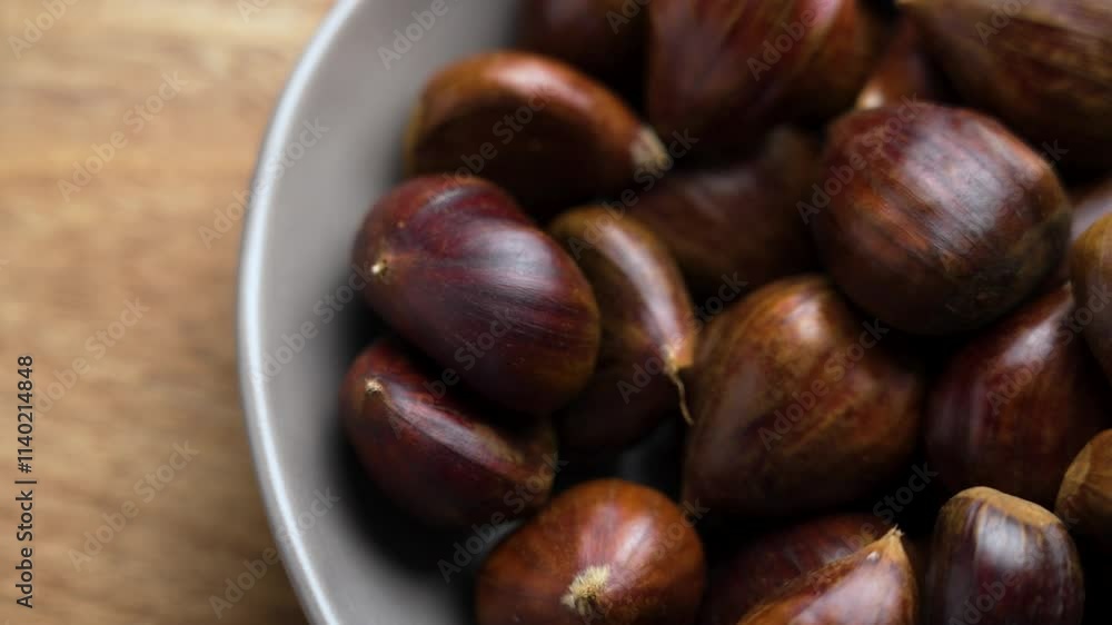 Whole unpeeled raw chestnut fruits with dark brown shells falling into a ceramic bowl on textured wooden table. Slow motion Whole unpeeled raw chestnut fruits with dark brown shells falling into a ceramic bowl on textured wooden table. Slow motion