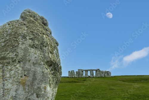 Close up of heel stone that indicatis location of solstice with in background Stonehenge prehistoric structure on Salisbury Plain, Wiltshire, England and moon in blue sky photo