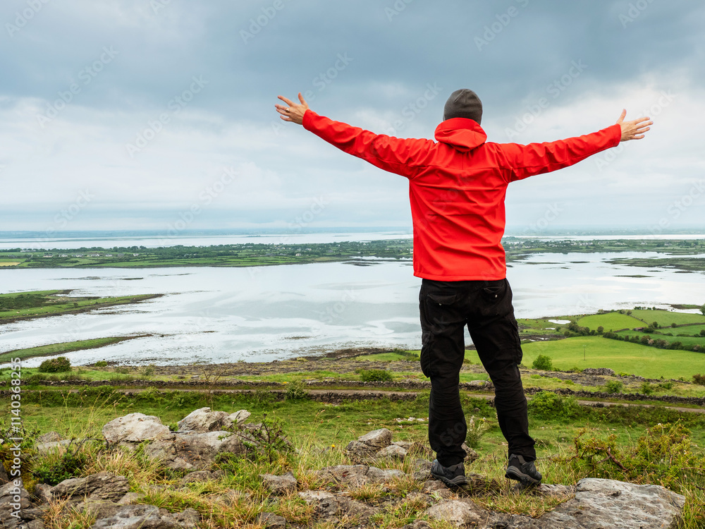 A man in a red jacket stands on a hill overlooking a valley and Galway bay. Male hands up in the air. Cloudy sky. Irish nature scene. Concept of travel and tourism. Burren, Ireland. A man in a red jacket stands on a hill overlooking a valley and Galway bay. Male hands up in the air. Cloudy sky. Irish nature scene. Concept of travel and tourism. Burren, Ireland.