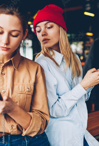 Hipster blogger spying online chat on smartphone of friend.Addicted best friends in casual outfit ignoring live communicating while spending leisure time in social networks on modern cellulars photo