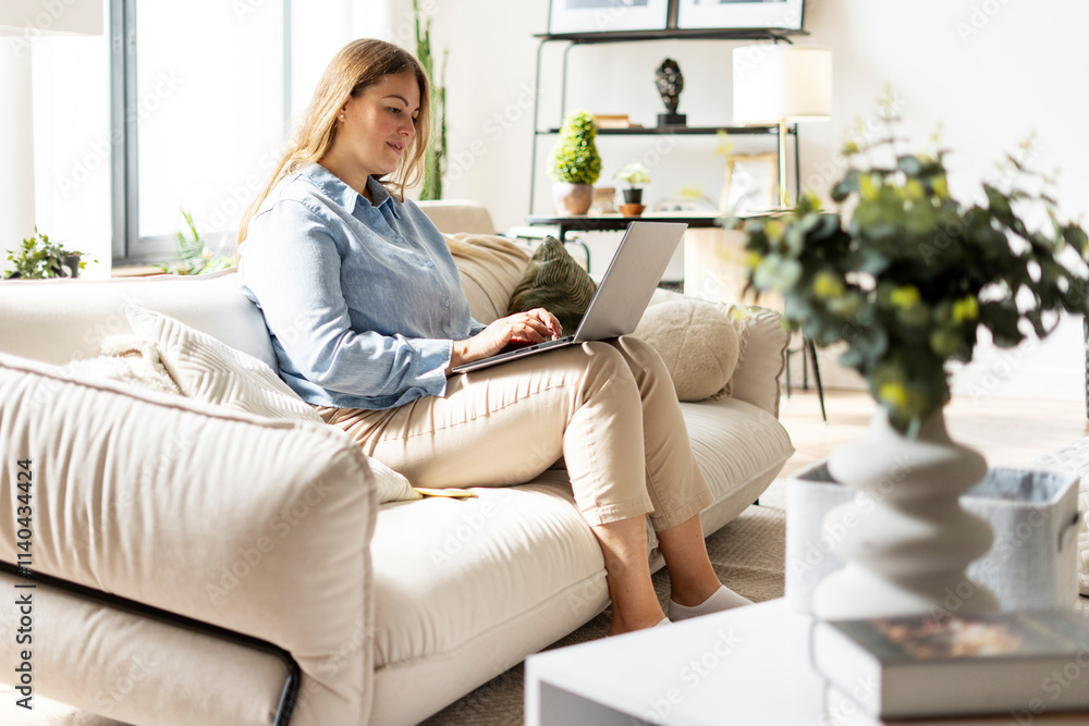 Smiling middle aged woman sitting on sofa and typing on laptop in living room at home Smiling middle aged woman sitting on sofa and typing on laptop in living room at home