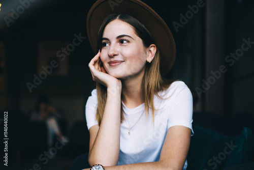 Smiling caucasian hipster girl in trendy hat and casual clothes sitting indoors enjoying free time, positive 20s young woman dressed in stylish apparel looking away dreaming about vacations #1140440022