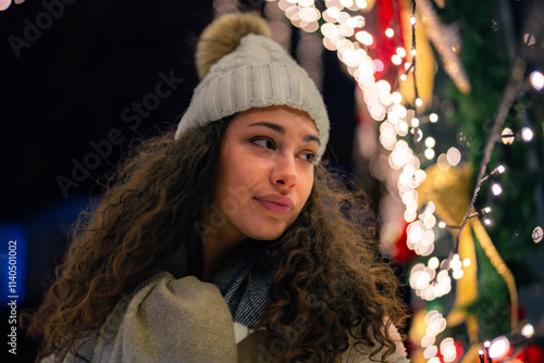 A woman in a winter hat and scarf stands beside festive holiday lights and decorations. The scene conveys a warm, cozy holiday atmosphere. Christmas concept.