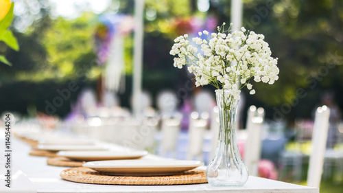 An elegantly set dining table at a wedding reception, featuring a pristine white plate, silverware, and a centerpiece of white flowers in a delicate vase, creating romantic and sophisticated ambiance