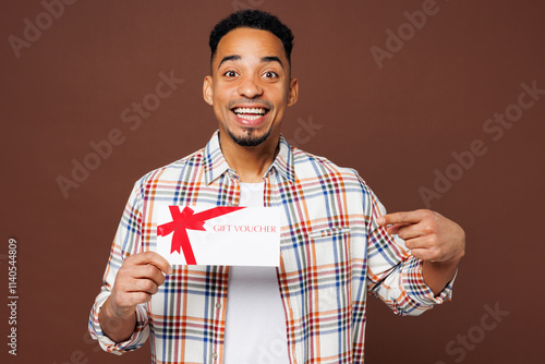 Young surprised shocked man of African American ethnicity he wears blue shirt casual clothes hold gift certificate coupon voucher card for store isolated on plain brown background. Lifestyle concept.