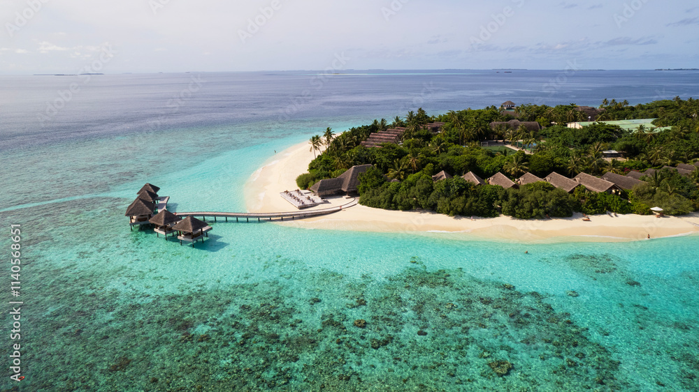 Turquoise clear water and the green island of Maldives. Top view of the ocean and the reef. Palm trees grow on the island. There are bungalows and villas. White sand on the beach. Fish are visible
