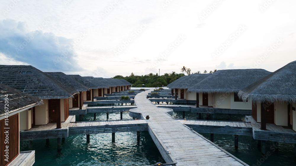 Bungalow on the water overlooking the green island. Thatched roofs, wooden bridge, water. Dark clouds. Green palm trees and a cell tower. A place to relax. Flowers grow in flower beds in places.
