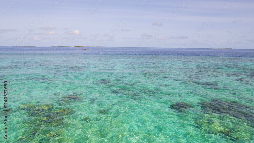 Turquoise clear ocean water. The reef is visible under the water. In the distance there is a hydroplane pad, islands and white clouds. The gradient of the water shimmers from light blue to dark