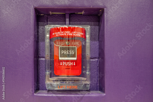 Red fire alarm system pull station under a protective clear plastic cover.  Fire alarm in a gymnasium mounted on purple wall with purple protective wall pads.