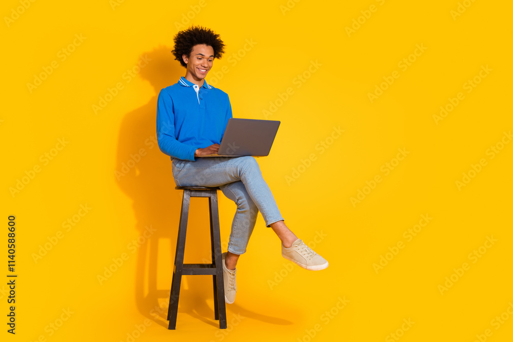 Full length photo of handsome young guy sit wooden chair netbook dressed stylish blue garment isolated on yellow color background Full length photo of handsome young guy sit wooden chair netbook dressed stylish blue garment isolated on yellow color background