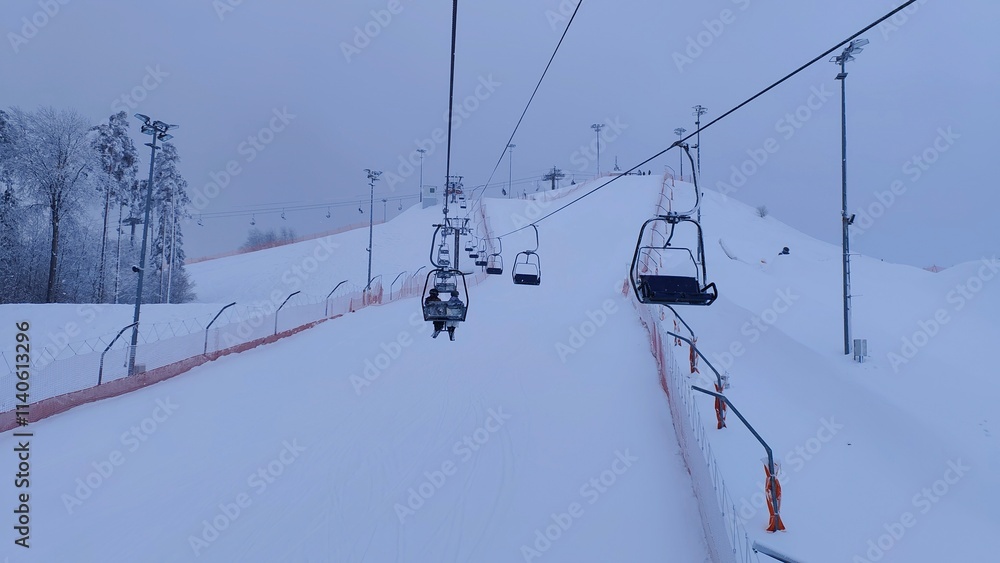 The snow-covered slopes with lampposts for skiers and snowboarders at the ski center are a mesh fence for safety and surrounded by a forest. Visitors go up the chairlift. Overcast and dense cloudiness The snow-covered slopes with lampposts for skiers and snowboarders at the ski center are a mesh fence for safety and surrounded by a forest. Visitors go up the chairlift. Overcast and dense cloudiness