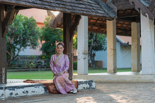 A young woman in a traditional Thai dress is peacefully sitting in a wooden temple pavilion. photo